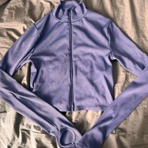 Blå ribbad zip-tröja - Säljer en blå ribbad tröja med dragkedja framtill och hög krage. Tröjan är croppad och har långa ärmar med tumhål, vilket ger en sportig och trendig vibe. Perfekt att styla med jeans eller kjol för en chill look.