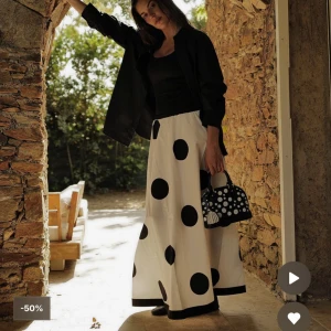 Vit långkjol med stora svarta prickar - Superfin långkjol i vitt med stora svarta prickar och bred svart kant längst ner. Kjolen har en luftig och flowy passform med resår i midjan. Perfekt för dig som vill sticka ut med en cool och lekfull stil.