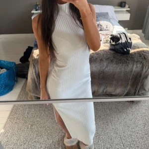 Vit ribbad långklänning med slits - Supersnygg vit ribbad långklänning med hög hals och slits på sidan. Ärmlös modell som sitter figurnära och ger en clean och fräsch look. 