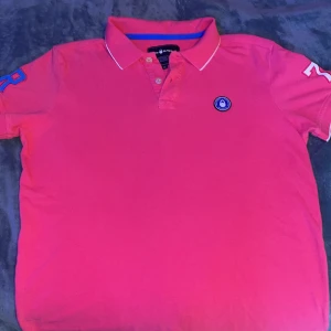 Rosa pikétröja från Sail Racing - Snygg rosa pikétröja från Sail Racing med klassisk krage, knappar och blå detaljer. Broderad logga på bröstet och tryck på kragen. Siffror på ärmarna ger en sportig vibe. Perfekt för dig som gillar färg och stilren design.