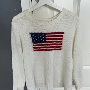 Vit stickad tröja med USA-flagga - Vit stickad tröja med rund hals och lång ärm. På bröstet finns en stor amerikansk flagga i blått, rött och vitt. Tröjan har ribbade muddar vid ärmslut och nederkant, perfekt för dig som gillar klassisk stil.