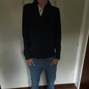 Svart långärmad pikétröja Tommy Hilfiger - Svart långärmad pikétröja från Tommy Hilfiger med klassisk krage och knappslå med randig detalj. Liten broderad logga på bröstet. Skön bomullskvalitet och stilren look som funkar till jeans eller chinos.