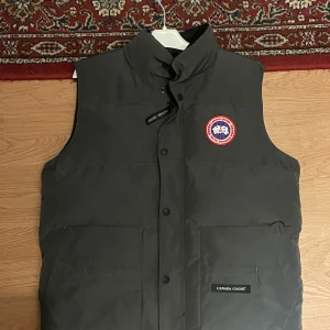 Svart dunväst från Canada Goose - Säljer en svart dunväst från Canada Goose med klassisk logga på bröstet. Västen har hög krage, stora fickor framtill och stängs med knappar. Perfekt för lager-på-lager och riktigt snygg till streetwear-stilen. Knappt använd.
