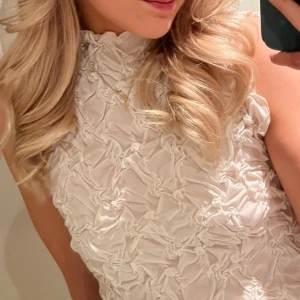 Vit croppad ärmlös topp med struktur - Supersnygg vit croppad topp med hög hals och ärmlös design. Tyget har en unik, skrynklig struktur som ger cool textur och extra attityd. Perfekt att styla med jeans eller kjol för en trendig look. Lite lätta sminkfläckar på (bild 4), går säkert bort med rätt medel!