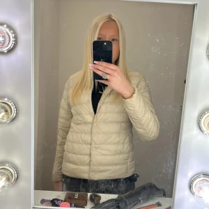 Beige dunjacka från Uniqlo  - Säljer en superlätt och smidig beige dunjacka från Uniqlo. Jackan har en clean look utan krage, tryckknappar framtill och quiltad design. Perfekt för lager-på-lager och enkel att ta med sig. Passar dig som gillar stilren och enkel vibe.