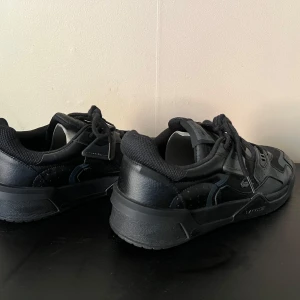 Svarta sneakers från Lacoste - Snygga svarta sneakers från Lacoste med coola detaljer och logga på sidan. Skorna har en mix av skinn och textil, samt snörning och platt sula. Perfekta för dig som gillar en sportig och stilren look.