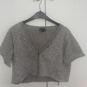 Glittrig grå croppad kofta KappAhl - Kort, glittrig grå kofta från KappAhl med v-ringning och knapp framtill. Koftan har korta ärmar och är croppad i modellen, perfekt att slänga över en topp för en extra touch. Stickad känsla och snyggt glittrande material.