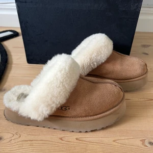 UGG tofflor i mocka och fårskinn - Mysiga UGG tofflor i beige mocka med fluffig vit fårskinnskant och innersula. Perfekta för kalla dagar hemma, med platt sula och rund tå. Klassisk UGG-logga på sidan och riktigt skön passform.