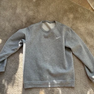 Grå sweatshirt från Nike - En klassisk grå sweatshirt från Nike med broderad vit swoosh på bröstet. Tröjan har rund halsringning, långa ärmar och ribbade muddar. Perfekt för en chill och sportig look. Materialet är mjuk bomullsmix som känns skön mot huden.