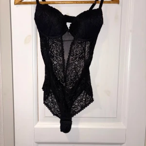 Spetsbody - Säljer en svart korsett med vackert blommigt spetsmönster och vadderade kupor. Axelbanden är justerbara och ryggen har en smidig knäppning. Tryckknappar i grenen. . Perfekt för dig som vill ha en snygg och feminin look med extra detaljer.