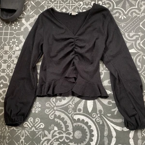 Svart långärmad blus med volang - Svart blus med v-ringning, rynkdetalj framtill och volangkant nedtill. Långa ärmar med lätt puff vid handlederna. ☀️