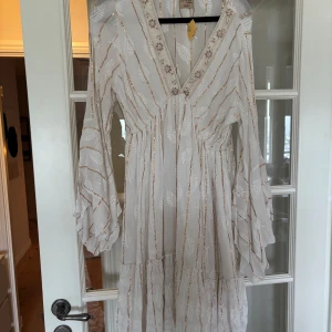 Vit boho-klänning med volangärm - Superfin vit kortklänning i boho-stil med guldiga ränder och broderade blad. Klänningen har v-ringning med spetsdetaljer och vida, volangiga långärmar. Köpt i Grekland, aldrig använd! Prislapp kvar. Storlek One Size men passar mig som har xs/s 