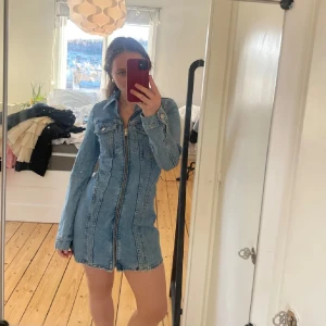 Kort jeansklänning med dragkedja - Säljer en snygg kort jeansklänning i ljusblå denim med långa ärmar och markerad midja. Klänningen har hel dragkedja framtill, klassisk skjortkrage och bröstfickor med knappar. Perfekt för dig som gillar en cool och trendig look.