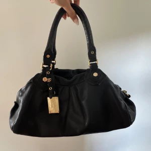 Svart Marc Jacobs väska med gulddetaljer - Säljer min Marc Jacobs väska som jag älskar men tyvärr inte kommer till användning av längre.  Skicket är mycket fint och har inga större defekter, obs att den har en liten fläck på insidan (se sista bilden) men annars är allt som nytt.  Extra band kommer med