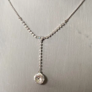 Glittrigt silverhalsband med strass - Ett elegant halsband i silverfärg med gnistrande strass-stenar och en droppformad hängande kristall. Kedjan har små, tätt sittande stenar och ett justerbart lås baktill. Perfekt för dig som vill ha något extra glittrigt och stilrent.
