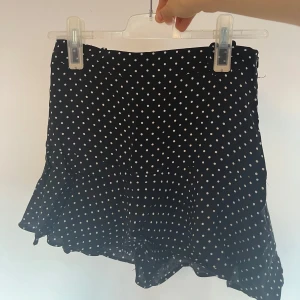 Svart prickig kortkjol med volang och inbyggda shorts! - Supersöt svart kortkjol med vita prickar och volangkant. Kjolen har en lätt och flowy känsla, perfekt för att skapa en lekfull look. Stängs med dragkedja i sidan och har en feminin passform som sitter snyggt i midjan.💞💞