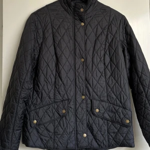 Svart quiltad jacka från Barbour - Quiltad svart jacka från Barbour med klassiskt rutmönster, guldiga tryckknappar och dragkedja. Jackan har två stora fickor framtill och hög krage. Perfekt för dig som gillar stilren och tidlös look. Materialet är polyester och jackan är långärmad.