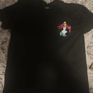 Svart Stüssy t-shirt med färggrafik - Svart t-shirt från Stüssy med stor, färgglad grafik och tryck på både fram- och baksida. Framsidan har Stüssy-logga och en staty med blomma, baksidan har collage med blommor, djur och text i rosa. Skön passform och cool streetwear vibe.