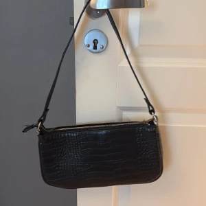 Svart axelväska  - Snygg svart axelväska med croco-mönstrad yta och dragkedja upptill. Stilren och enkel design med axelremsband. 