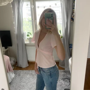 Ljusrosa omlottopp med knytband - Supersöt ljusrosa omlottopp med djup v-ringning och korta, lite fladdriga ärmar. Toppen har knytband i midjan som knyts i ryggen för en snygg passform. Perfekt till jeans eller kjol för en fräsch look.