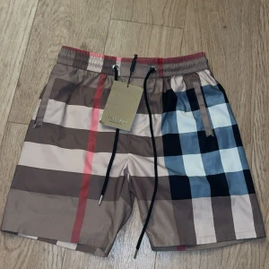 Rutig Burberry shorts med snörning - Snygga shorts från Burberry med klassiskt rutigt mönster i beige, brunt, svart, vitt och blått. De har elastisk midja med svart snörning och praktiska fickor. Perfekta för sommaren och ger en lyxig vibe till din outfit.