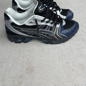 ASICS gel kayano 14  - Snygga Asics sneakers i svart, silver och mörkblå med coola detaljer och meshpaneler för extra andningsförmåga. Skorna har snörning och en chunky sula som ger grym komfort. Perfekta för dig som gillar sportig stil och vill sticka ut.