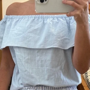 Ljusblå offshoulder topp med volang - Superfin ljusblå offshoulder topp med bred volang runt hela axelpartiet. Luftig och lätt i bomullsmaterial, perfekt för varma dagar. Enkel och stilren design som ger en fräsch look.