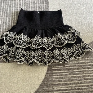 Svart volangkjol med broderad spets - Supersöt svart kortkjol med bred smock i midjan och flera lager volanger. Kjolen har vit broderad spets längs volangkanterna som ger en unik och cool look. Perfekt för dig som gillar att sticka ut med detaljer och volym.
