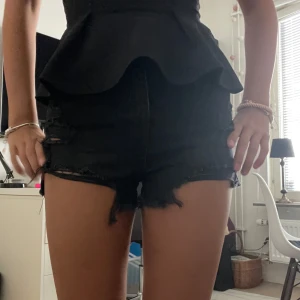 Svarta jeansshorts med fransig kant - Snygga svarta jeansshorts med hög midja och slitna detaljer framtill. Shortsen har rå, fransig kant nertill och klassiska bakfickor. Perfekta för sommaren och ger en cool vibe till din outfit.