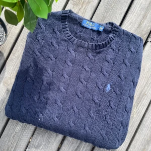 Mörkblå kabelstickad tröja Polo Ralph Lauren - Kabelstickad mörkblå tröja från Polo Ralph Lauren med rund halsringning och broderad logga på bröstet. Tröjan är långärmad och har ribbade muddar vid ärmslut och nederkant. Perfekt för lager-på-lager under hösten och vintern.