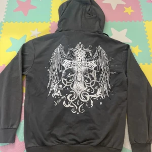 Grå hoodie med kors och vingar - Mörkgrå hoodie med dragkedja och huva, från Konwei. På ryggen finns ett stort tryck med kors och vingar i vitt och grått, samt mindre kors på bröstet. Tröjan har långa ärmar, snörning i huvan och känguruficka framtill. 