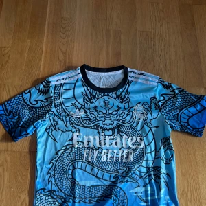 Real Madrid x Adidas Bellingham tröja M - Snygg Real Madrid fotbollströja från Adidas i blått med svart drakmönster och vita detaljer. Kortärmad modell med svart krage, klubbmärke och sponsortryck framtill samt Bellingham och nummer 5 på ryggen. Tillverkad i lätt och ventilerande polyester.