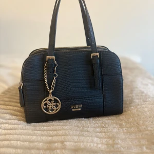 Svart handväska från Guess med gulddetaljer - Snygg svart handväska från Guess med strukturmönster och guldfärgad logga samt kedjehänge. Väskan har dubbla handtag, dragkedja upptill och flera fack inuti. Perfekt storlek för det viktigaste och ger en lyxig känsla till din outfit.