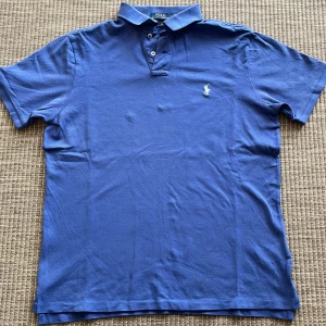 Blå pikétröja från Polo Ralph Lauren - Snygg blå pikétröja från Polo Ralph Lauren i bomull. Klassisk krage, korta ärmar och broderad logga på bröstet. Modellen är custom slim fit och passar perfekt till en avslappnad men stilren look.