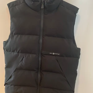 Svart dunväst från Sail Racing - Svart dunväst från Sail Racing med hög krage och dragkedja framtill. Västen har två snedställda fickor och diskret logga på bröstet. Tillverkad i syntetmaterial med Primaloft-fyllning som håller värmen. Perfekt för lager-på-lager under kyliga dagar. Köpt för 4000kr