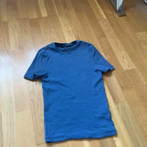 Blå ribbad t-shirt från BikBok - En basic blå t-shirt från BikBok i ribbad bomullskvalitet. Modellen är klassisk med rund hals och korta ärmar, perfekt till jeans eller kjol. Enkel och stilren design som funkar till det mesta.
