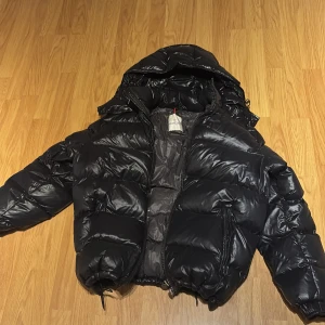 Svart Moncler pufferjacka med huva - Säljer en svart pufferjacka från Moncler med glansig finish och huva. Jackan har dragkedja framtill, två stora fickor med dragkedja och ikonisk Moncler-logga på ärmen. Insidan har ett coolt serietidningsmärke. Perfekt för kalla dagar och riktigt snygg streetstil. (Priset är inte hugget i sten, hör av er vid minsta fundering)😁🫡