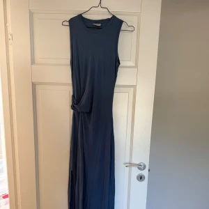 Blå ärmlös långklänning med knut - Snygg blå långklänning i mjukt material med ärmlös design och cool knutdetalj i midjan från By Malene Birger. Klänningen har en slits på sidan som ger extra rörelse. Perfekt för dig som gillar stilrena och mjuka plagg! Pris kan diskuteras.