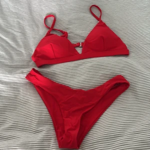 Röd trekantsbikini med spänne - Snygg röd bikini med trekantsformad topp och smala axelband. Toppen har ett metallspänne i ryggen och lätt vadderade kupor. Bikinitrosan är enkel och högt skuren. Perfekt för stranden eller poolen i sommar. (Har använt dem en gång)