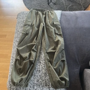 Olivgröna cargobyxor från Shein - Säljer ett par olivgröna cargobyxor från Shein med elastisk midja och muddar vid bensluten. Byxorna har fickor på sidorna och är tillverkade i ett lätt syntetmaterial. Perfekta för en avslappnad och trendig look använd en gång.