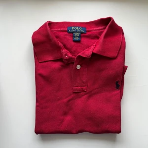 Röd pikétröja från Ralph Lauren - Klassisk röd pikétröja från Polo Ralph Lauren med korta ärmar och broderad marinblå logga på bröstet. Tröjan har krage, två knappar och är tillverkad i bomull. Perfekt för en stilren och avslappnad look.