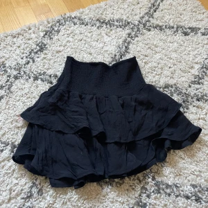 Svart volangkjol med hög midja - Supersöt svart kortkjol med volanger och hög smockad midja. Kjolen har en flowy känsla och passar perfekt till sneakers eller boots. Materialet känns lätt och luftigt, perfekt för varmare dagar.