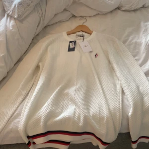 Vit Moncler tröja med ränder - Vit långärmad tröja från Moncler med strukturerad yta och broderad logga på bröstet. Ribbstickade muddar och nederkant med röd, blå och vit rand. Klassisk och stilren design som passar perfekt till jeans eller chinos.