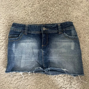 Vintage kort lågmidjad jeanskjol - Säljer en snygg mörkblå jeanskjol med slitningar. Vet tyvärr inte märket. Måtten står i bilderna. Kjolen är knappt använd och är lågmidjad och kort. 