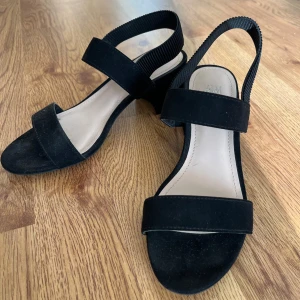 H&M svarta sandaletter 36 - Eleganta svarta sandaletter från H&M i storlek 36. Mockaimmitation. Remmar över foten och ribbad resår runt hälen. Foder och innersulor i läderimmitation  samt gummisulor. Perfekta för både vardag och fest. Bekväm modell med resårband. Klackhöjd 6.5 cm. I fint skick.