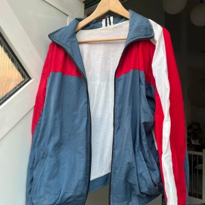 Retro vindjacka i rött, blått och vitt - Snygg retro vindjacka med färgblock i rött, blått och vitt. Jackan har dragkedja framtill, ståkrage och elastiska muddar. Perfekt för dig som gillar old school vibes och sportig stil. Fickor med dragkedja på sidorna och klassisk passform. Den är köpt på en marknad i Spanien så vet ej pris eller storlek men är lite stor på mig som vanligtvis bär s/m! Den har en liten fläck på ryggen annars har jag inte sett någon annan fläck🥰 