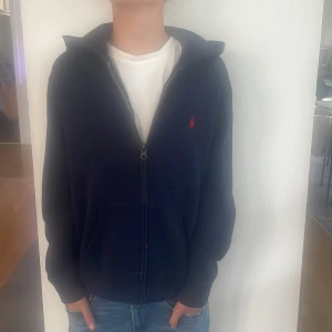 Mörkblå hoodiejacka från Polo Ralph Lauren - Mörkblå hoodiejacka från Polo Ralph Lauren med dragkedja och huva. Klassisk röd logga broderad på bröstet och kängurufickor framtill. Skön och avslappnad passform, perfekt till jeans och t-shirt. Tröjan har är änvand men är fof efter bra tvätt ungefär som ny! Nypris ungefär 1200 kr, säljs för endast 350kr! Har inte kvitto men är köpt på kidsbrandstore i Nacka forum, känner du dig osäker så ställ frågor. Tack för din tid!