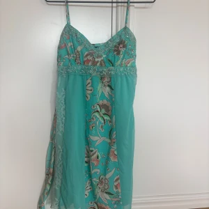 Turkos blommig kortklänning från Triangles - Superfin turkos kortklänning från Triangles med blommigt mönster, tunna axelband och spetsdetaljer. Klänningen har omlottkänsla.
