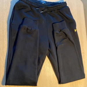 Svarta mjukisbyxor Lyle & Scott - Svarta mjukisbyxor från Lyle & Scott med elastisk midja och snörning. Byxorna har ribbade muddar vid bensluten, sidofickor och en bakficka. Liten gul logga broderad på ena benet. Perfekta för chill eller träning.