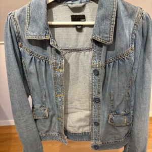 Ljusblå jeansjacka från H&M - Snygg ljusblå jeansjacka från H&M med klassisk krage, knappar framtill och två fickor med lock. Jackan har rynkade detaljer vid axlar och bröst, vilket ger en cool och trendig look.💗